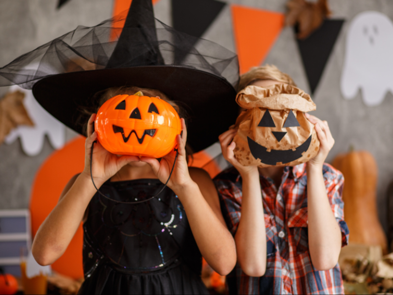 Two children holding pumpkin masks in front of their faces at a Halloween‑themed setting with decorations.