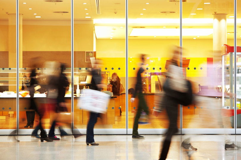 People walking past a glass‑walled café with warm yellow lighting, shown in motion blur inside a busy indoor space.
